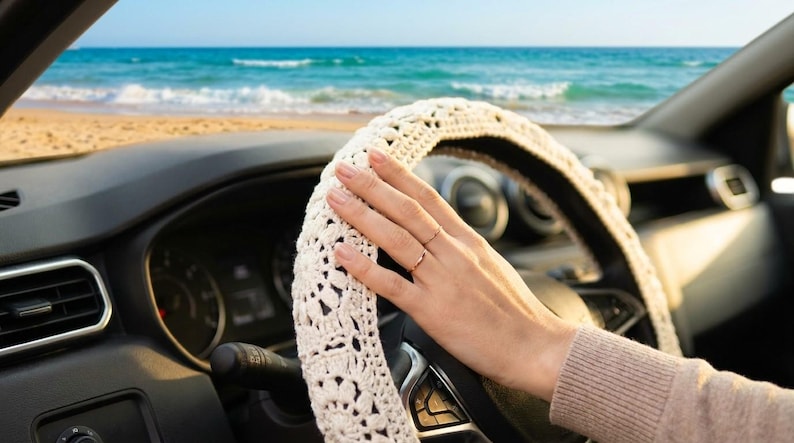 A ivory steering wheel cover is shown in a car with a beach and ocean in the background. The cover has an open, lacy design. The car's dashboard and steering wheel are black.