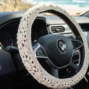 Puede incluir: Funda de volante de ganchillo color crema. La funda est&aacute; hecha de un hilo suave y texturizado y presenta un patr&oacute;n floral. La funda est&aacute; instalada en un volante negro en un coche.