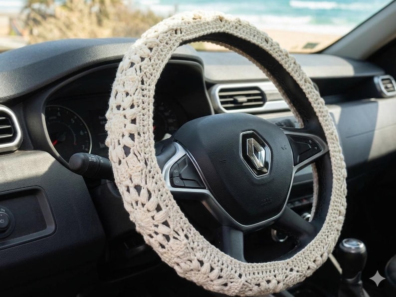 Puede incluir: Funda de volante de ganchillo color crema. La funda est&aacute; hecha de un hilo suave y texturizado y presenta un patr&oacute;n floral. La funda est&aacute; instalada en un volante negro en un coche.