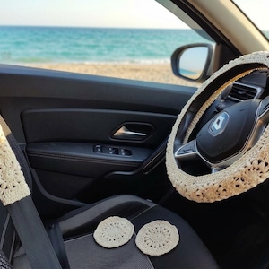 Beige crochet steering wheel cover, seatbelt cover, and two coasters inside a car. The car interior is black with a view of the ocean and beach in the background. The crochet items have a floral design.