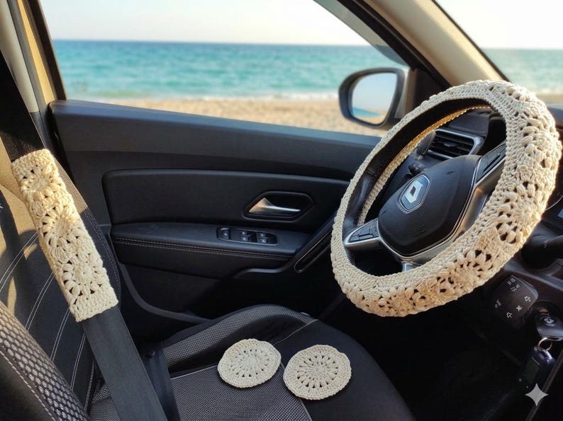 Beige crochet steering wheel cover, seatbelt cover, and two coasters inside a car. The car interior is black with a view of the ocean and beach in the background. The crochet items have a floral design.