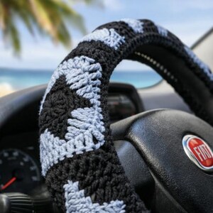 May include: A black and light blue crocheted steering wheel cover. The cover is made of yarn and has a geometric pattern. The steering wheel is black and has a red and silver Fiat logo.