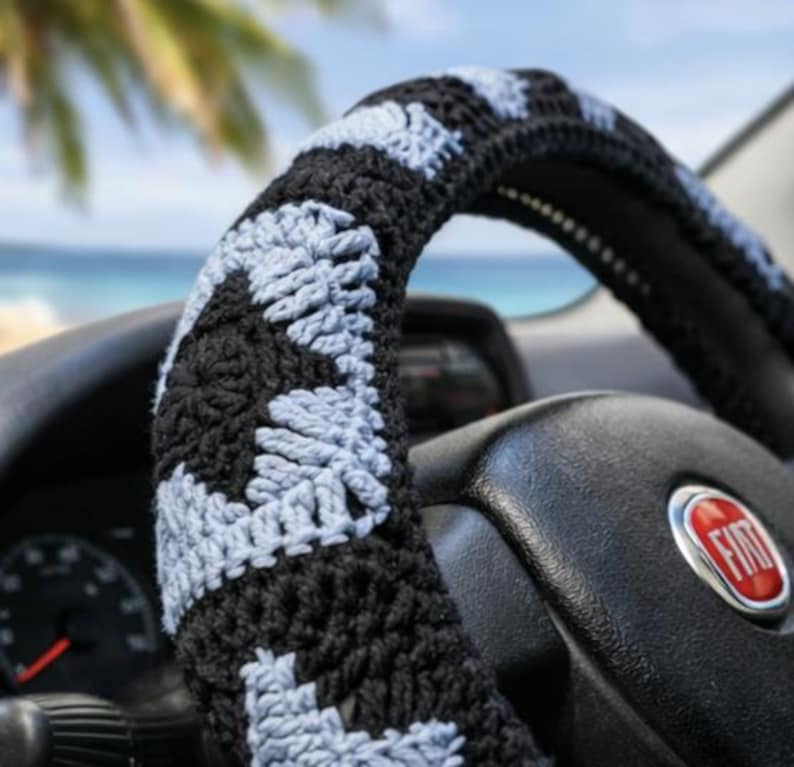 May include: A black and light blue crocheted steering wheel cover. The cover is made of yarn and has a geometric pattern. The steering wheel is black and has a red and silver Fiat logo.