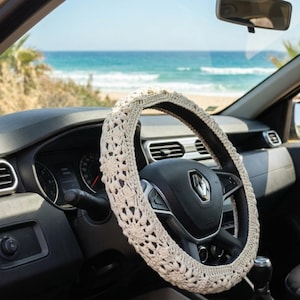 May include: A cream-colored, crocheted steering wheel cover with an intricate, openwork floral pattern. The cover is fitted snugly over a black steering wheel inside a vehicle, featuring a central Renault logo on the horn button.