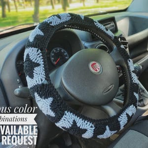 A black and gray steering wheel cover with star patterns. The cover is on a black steering wheel inside a car. Text on the image reads "Various color combinations ARE AVAILABLE UPON REQUEST."