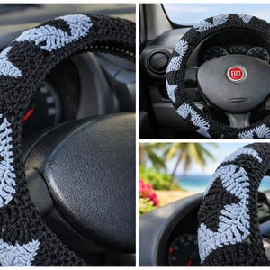 May include: Black and light blue crocheted steering wheel cover. The cover features a repeating pattern of light blue shapes on a black background. The cover is shown on a car's steering wheel.