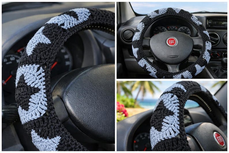 May include: Black and light blue crocheted steering wheel cover. The cover features a repeating pattern of light blue shapes on a black background. The cover is shown on a car's steering wheel.