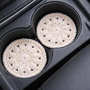 Two cream-colored, crocheted coasters with a floral design sit in a car's cup holders. The coasters are round and made of a textured yarn. The cup holders are black, and the car's interior is visible in the background.