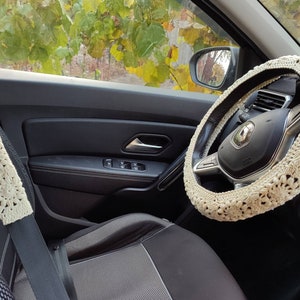 Puede incluir: Juego de fundas de ganchillo color crema para volante y cintur&oacute;n de seguridad. Las fundas presentan un patr&oacute;n floral y est&aacute;n instaladas en un coche. El interior del coche es negro y gris, con una vista del follaje fuera de la ventana.