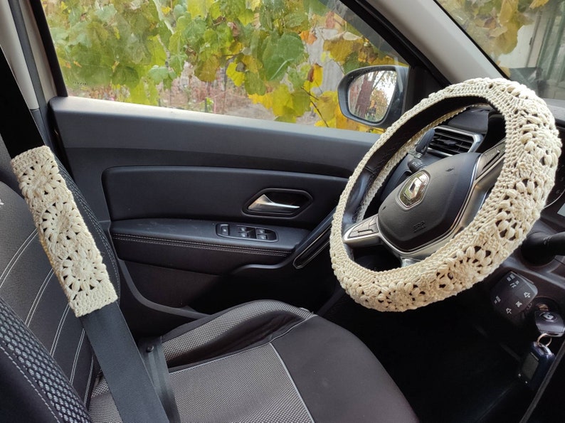 Puede incluir: Juego de fundas de ganchillo color crema para volante y cintur&oacute;n de seguridad. Las fundas presentan un patr&oacute;n floral y est&aacute;n instaladas en un coche. El interior del coche es negro y gris, con una vista del follaje fuera de la ventana.