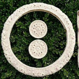 A cream-colored crochet steering wheel cover, two armrest covers, and two matching coasters. The armrest covers have wooden buttons. The set is displayed on a bed of green foliage.