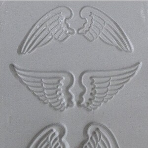 May include: Three different sized angel wing designs are carved into a gray textured surface. The wings are detailed with lines and curves.