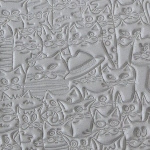 Op de afbeelding: Een close-up van een grijs, getextureerd oppervlak met een herhalend patroon van geëmbosseerde katten. Sommige katten dragen hoeden.