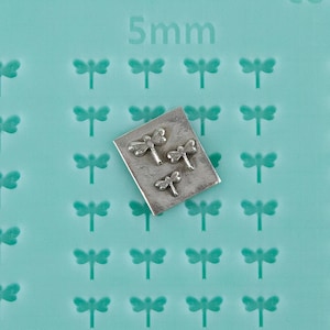 May include: A close-up shot of a teal-colored surface with a repeating dragonfly pattern and the text "5mm." A small, rectangular metal plate holds three silver dragonfly charms. The charms are detailed with wings and bodies.