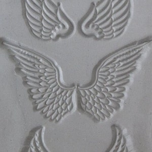 May include: A close-up of a silicone mold with four different angel wing designs. The mold is gray and has a textured surface.
