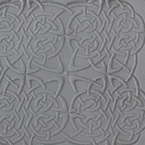 Może przedstawiać: Szara, kwadratowa, dekoracyjna płytka z wzorem celtyckiego węzła. Wzór jest wypukły i fakturowany, tworząc efekt 3D.