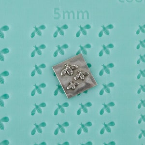 Puede incluir: Placa metálica cuadrada con tres pequeños dijes de abeja. Sobre una superficie azul claro con muchos diseños de abejas en relieve. El número "5mm" está impreso en la superficie.