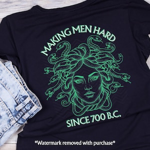 May include: Black t-shirt featuring a green graphic of Medusa's face and the text "MAKING MEN HARD SINCE 700 B.C." The design shows Medusa's face with snakes for hair. The shirt is folded with a pair of blue jeans.