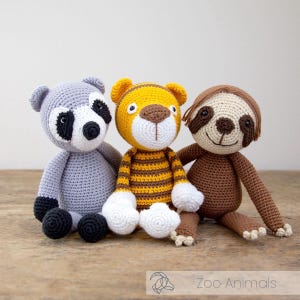 May include: Three handmade crochet zoo animal toys: a gray raccoon with black accents, a yellow and brown tiger, and a brown sloth. The toys are sitting on a wooden surface. The text "Zoo Animals" is at the bottom.