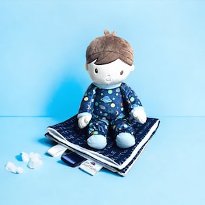 May include: A plush doll with brown hair and a blue and white space-themed pajama set. The doll sits on a navy blue blanket with white trim and the text "Twinkle Little Star". The blanket has satin tags and a "Sweet Cheeks" label.