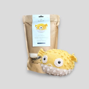 May include: A knitting kit for making a yellow and white pufferfish. The kit includes yarn, needles, and instructions. The kit is packaged in a brown paper bag with a label that says "Knitting Kit: Baby Blowfish".