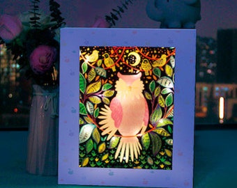 May include: A light blue wooden frame with a white border surrounds a colorful illustration of an owl with a moon and stars in the background. The owl is surrounded by green leaves and flowers. The frame is lit from behind, creating a glowing effect.