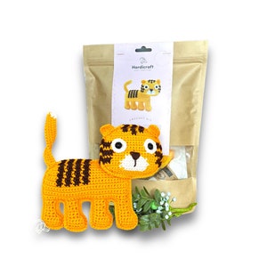 May include: A crochet tiger kit in a brown paper bag. The kit includes yarn, stuffing, and instructions to make a crocheted tiger toy. The finished tiger is orange with black stripes and white accents.