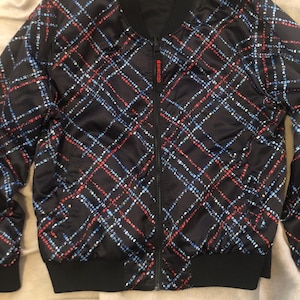May include: A black bomber jacket featuring a diagonal plaid pattern composed of small red, white, and blue icons. The jacket has a black ribbed collar, cuffs, and hem, a central zipper closure, and a red zipper pull tab with the text WWDC20.