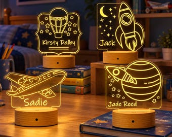 May include: Four illuminated acrylic night lights with wooden bases, featuring engraved space-themed designs and names: Kirsty Dalby with a hot air balloon, Jack with a rocket, Sadie with an airplane, and Jade Reed with a planet and rocket.