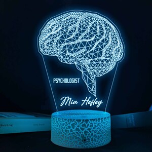 Può includere: Una luce notturna a forma di cervello blu stampata in 3D con il testo "Psicologa Mia Hefley" sulla base.