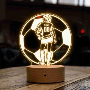 May include: A wooden base with a light up 3D design of a soccer player holding a soccer ball. The design is outlined in yellow and the player is wearing a jersey with the text "NAME 00".