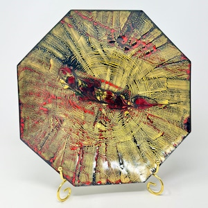 May include: An octagonal decorative plate with a gold, red, and black abstract design. The plate is supported by a gold-colored metal stand. The plate's design features a radial pattern with a central, elongated red and black element.