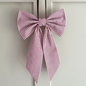 May include: A decorative fabric bow featuring a vertical striped pattern in deep maroon and white. The bow is constructed with two large, pleated upper loops and two long, pointed hanging tails, suspended by a thin dark ribbon loop at the top.