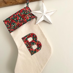 May include: A cream-coloured Christmas stocking featuring a cuff and a large letter B applique in a festive red berry and green leaf floral pattern. The stocking includes a matching fabric loop for hanging and is displayed with a white star ornament.