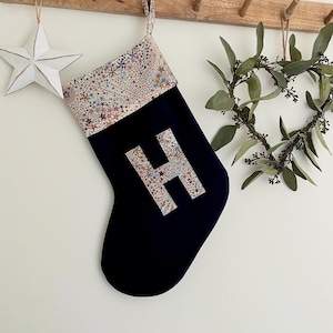 May include: A navy blue Christmas stocking featuring a cuff and a large letter H applique, both made from a light-coloured fabric with a colourful pattern of small stars, moons, and celestial shapes. The stocking includes a matching fabric hanging loop.