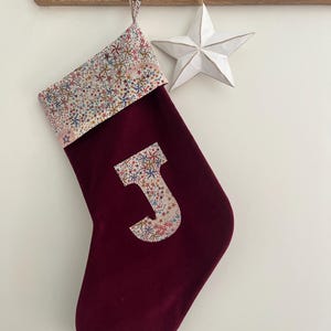 Op de afbeelding: Een bordeauxrode kerstsok met een patroonmanchet en een letter "J" applicatie. De manchet en de letter hebben een sterrenontwerp. Een witte sterdecoratie hangt naast de sok, wat een feestelijke uitstraling creëert.