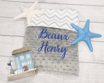 May include: Gray and white chevron baby blanket with a gray minky dot texture. The blanket has the name "Beaux Henry" embroidered in blue thread. The blanket is surrounded by two starfish, one white and one blue.