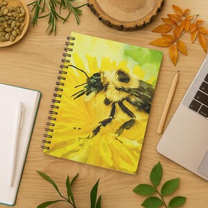 Może przedstawiać: Notatnik ze spiralą z ilustracją w stylu akwareli przedstawiającą puszystego trzmiela odpoczywającego na dużym, jaskrawożółtym kwiecie. Okładka prezentuje odcienie jasnego żółtego, zieleni i czerni z czarną spiralną oprawą.