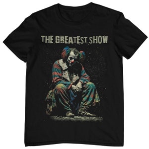 Könnte beinhalten: Schwarzes T-Shirt mit einem Aufdruck eines gruseligen Clowns, der sitzt. Der Text "THE GREATEST SHOW" ist über dem Clown gedruckt.