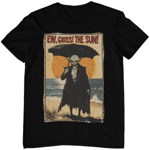Könnte beinhalten: Ein schwarzes T-Shirt mit einer Grafik im Vintage-Stil, die einen Vampir zeigt, der einen Regenschirm hält und an einem Strand entlangläuft. Die Grafik lautet "Ew, Gross! The Sun!"