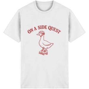 Könnte beinhalten: Weißes T-Shirt mit roter Grafik einer Gans mit Helm und Rollschuhen, darüber der Text "ON A SIDE QUEST". Das Design ist mittig auf der Vorderseite des Shirts platziert.