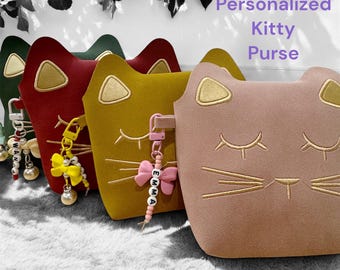 May include: Three personalized kitty purses in red, yellow, and pink with embroidered cat faces and gold accents. Each purse has a detachable charm with a bow and a beaded name.