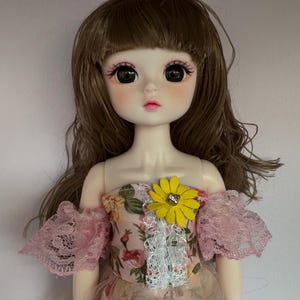May include: A doll with brown hair and a floral dress. The doll has large, dark eyes and a pink blush on her cheeks. She is wearing a pink and white lace off-the-shoulder dress with a yellow flower brooch.