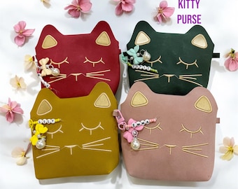 May include: Four personalized kitty purses in different colors: red, green, yellow, and pink. Each purse has a cat face with closed eyes and a gold embroidered nose and whiskers. Each purse has a charm with a name on it: Lily, Sophie, Emma, and another name that is not visible.