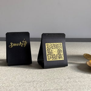 Puede incluir: Dos expositores triangulares negros con detalles dorados. Un expositor tiene la palabra "Smokey" en letras doradas. El otro expositor tiene un código QR dorado.