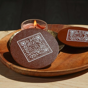 Puede incluir: Dos posavasos marrones con un diseño de código QR y las letras "GR" en blanco se muestran en una bandeja de madera. Una vela encendida en un portavelas de cristal está cerca. Los posavasos son redondos y parecen estar hechos de cuero o material similar.