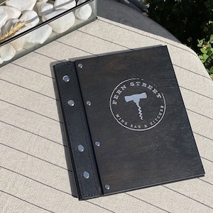 May include: A black wooden menu with the text "FERN STREET WINE BAR & KITCHEN" and a silver corkscrew logo. The menu is bound with a black leather spine and silver rivets. The menu is on a light-colored table.