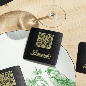 May include: A restaurant table setting featuring a decorative plate with a green bird and floral design, black coasters with QR codes and the "Banchetto" logo, and a stemmed glass with a gold rim.