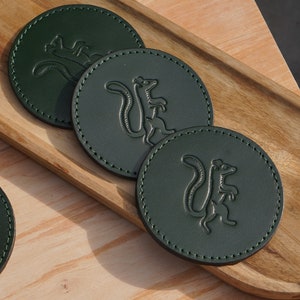 May include: Three dark green leather coasters with a stitched edge, each featuring an embossed image of a squirrel. The coasters are arranged on a wooden tray.