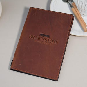 May include: A brown leather menu cover with the embossed text "BROWNSTONE RESTAURANT". The cover is rectangular with a stitched border. A white plate, silverware, and a glass are in the background.